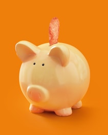 A ceramic piggy bank with a glossy finish stands against an orange background. Emerging from the slot is a single potato chip.