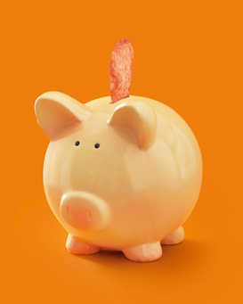 A ceramic piggy bank with a glossy finish stands against an orange background. Emerging from the slot is a single potato chip.