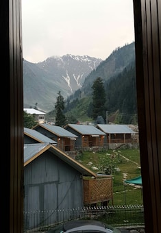 A mountain landscape with several small cabins or houses in the foreground. The cabins have metal roofs and wooden walls, surrounded by greenery. In the background, snow-capped mountains rise with dense forest covering the lower slopes. The scene is framed by a wooden structure, possibly a window or a doorway.