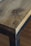 A close-up of a wooden table or surface with a natural finish, featuring distinct wood grain and a logo or branding mark burned into the corner. The edge of the surface is complemented by a metal frame, adding a rustic or industrial touch.