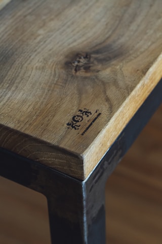A close-up of a wooden table or surface with a natural finish, featuring distinct wood grain and a logo or branding mark burned into the corner. The edge of the surface is complemented by a metal frame, adding a rustic or industrial touch.
