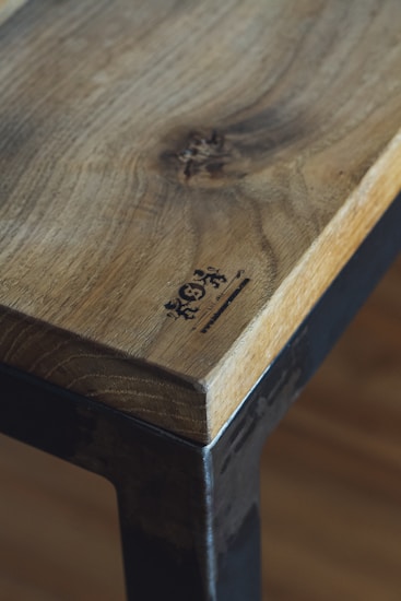 A close-up of a wooden table or surface with a natural finish, featuring distinct wood grain and a logo or branding mark burned into the corner. The edge of the surface is complemented by a metal frame, adding a rustic or industrial touch.