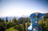 A bright blue binocular telescope is positioned on a scenic mountain viewpoint, overlooking lush green trees and distant mountain ranges under a clear blue sky.