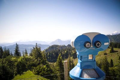 A bright blue binocular telescope is positioned on a scenic mountain viewpoint, overlooking lush green trees and distant mountain ranges under a clear blue sky.