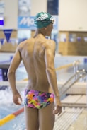 A swimmer wearing a colorful, patterned swimsuit stands by the edge of an indoor swimming pool. The person is wearing a green swim cap and appears to be ready for swimming. The pool area has blue and white decorations, with a water surface that is slightly rippled. The surroundings indicate an indoor competitive swimming facility.