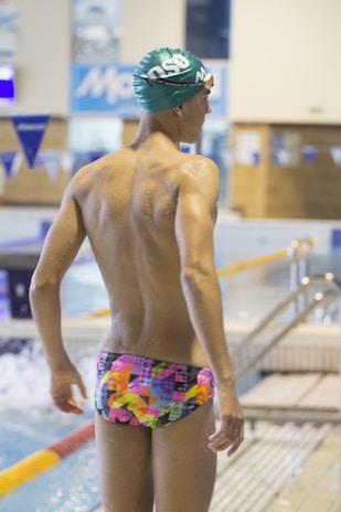 A swimmer wearing a colorful, patterned swimsuit stands by the edge of an indoor swimming pool. The person is wearing a green swim cap and appears to be ready for swimming. The pool area has blue and white decorations, with a water surface that is slightly rippled. The surroundings indicate an indoor competitive swimming facility.