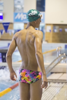 A swimmer wearing a colorful, patterned swimsuit stands by the edge of an indoor swimming pool. The person is wearing a green swim cap and appears to be ready for swimming. The pool area has blue and white decorations, with a water surface that is slightly rippled. The surroundings indicate an indoor competitive swimming facility.