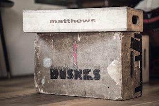 Two weathered wooden boxes are stacked on a wooden floor. The top box is smaller, light-colored, and marked with the word 'matthews'. The larger box beneath it is more worn, with visible marks, scratches, and the word 'BUSHES' printed on it. Both boxes have visible side handles.