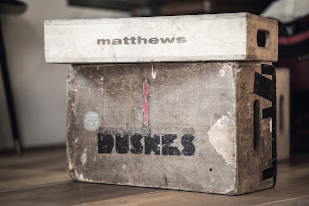 Two weathered wooden boxes are stacked on a wooden floor. The top box is smaller, light-colored, and marked with the word 'matthews'. The larger box beneath it is more worn, with visible marks, scratches, and the word 'BUSHES' printed on it. Both boxes have visible side handles.