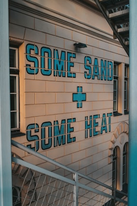 A building facade features bold blue letters on a beige wall that read 'SOME SAND + SOME HEAT'. The text is displayed in a block style, catching the light with a slightly industrial look. Metal stairs are visible in the foreground, adding an urban element to the scene.
