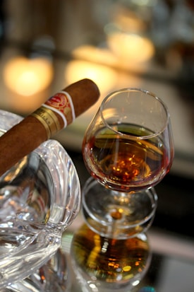 A glass of amber liquid, likely whiskey or brandy, sits on a reflective surface alongside a brown cigar placed on an elegant crystal ashtray. The setting suggests a luxurious and relaxed ambiance.