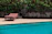 A sunlit wicker pool lounger beside a sparkling blue pool with lush greenery around.