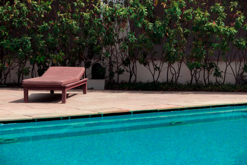 A cozy wicker pool lounger with soft cushions beside a shimmering blue pool under dappled sunlight.