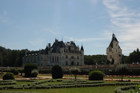 A grand historic chateau surrounded by manicured gardens and lush greenery under a clear blue sky. The architecture features pointed towers and classic stonework.