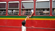 A red bus with yellow and green stripes is parked with several passengers visible through the windows. A person inside the bus reaches out through the window to receive a pink cup from a child wearing a black hat and white shirt standing outside.