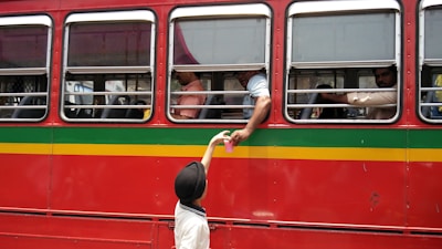 A red bus with yellow and green stripes is parked with several passengers visible through the windows. A person inside the bus reaches out through the window to receive a pink cup from a child wearing a black hat and white shirt standing outside.