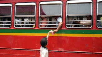 A red bus with yellow and green stripes is parked with several passengers visible through the windows. A person inside the bus reaches out through the window to receive a pink cup from a child wearing a black hat and white shirt standing outside.