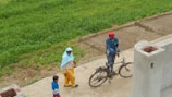 A man in a red turban and blue jacket stands next to an old bicycle on a concrete path. A woman wearing a blue headscarf and yellow pants walks along with a child in a blue sweater on the same path. The background includes a green grassy field and a partly-constructed brick wall.