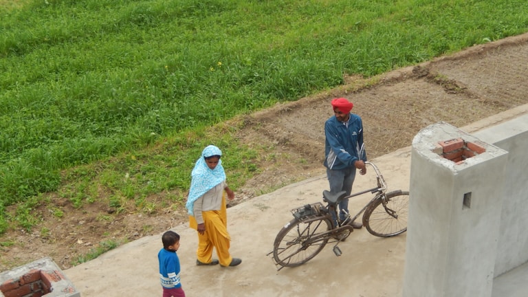 A man in a red turban and blue jacket stands next to an old bicycle on a concrete path. A woman wearing a blue headscarf and yellow pants walks along with a child in a blue sweater on the same path. The background includes a green grassy field and a partly-constructed brick wall.