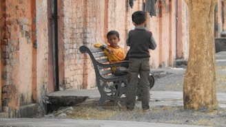 Two children are in a courtyard next to a brick wall. One child is sitting on a metal bench wearing a yellow sweater and looking toward the camera. The other child is standing and facing away from the camera. A tree is visible on the right side.