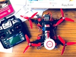 A drone with red propellers is placed on a wooden surface next to a remote control labeled 'FLYSKY'. Various documents and a book on mechanical engineering are scattered around. The drone appears to be in the process of assembly, with visible wiring and components.