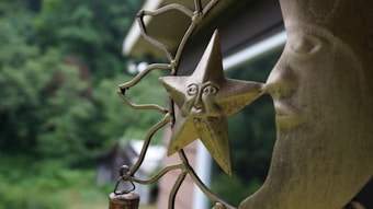 A decorative metal sculpture featuring a star with a facial design, surrounded by swirling metalwork, is set against a blurred greenery background. The image conveys a natural, outdoor setting.