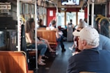 A group of people is seated inside a bus or tram. The interior features wooden seats and metal poles. The surroundings suggest a daytime setting with natural light coming through the windows. A digital display screen is located at the front, and there are hanging handles for standing passengers.