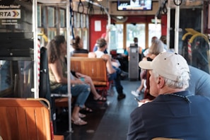 A group of people is seated inside a bus or tram. The interior features wooden seats and metal poles. The surroundings suggest a daytime setting with natural light coming through the windows. A digital display screen is located at the front, and there are hanging handles for standing passengers.