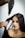 A woman with straight, dark hair is having her hair styled using a hairdryer and a round brush. The hand holding the brush appears to be styling her hair while she sits calmly, wearing a black top.