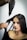 A woman with straight, dark hair is having her hair styled using a hairdryer and a round brush. The hand holding the brush appears to be styling her hair while she sits calmly, wearing a black top.