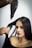 A woman with straight, dark hair is having her hair styled using a hairdryer and a round brush. The hand holding the brush appears to be styling her hair while she sits calmly, wearing a black top.