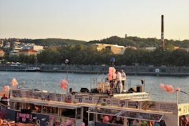A river barge decorated with pink balloons sails along a waterfront lined with trees and buildings. People are gathered on the barge, some standing on the deck under microphones and lights. The background features a green hillside and a tall smokestack.