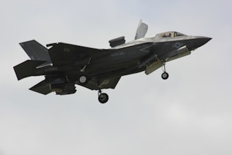 A military fighter jet is captured mid-flight against a pale sky. The aircraft features angular, stealthy design and dark gray color with visible landing gear and some markings on its body.
