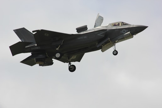 A military fighter jet is captured mid-flight against a pale sky. The aircraft features angular, stealthy design and dark gray color with visible landing gear and some markings on its body.