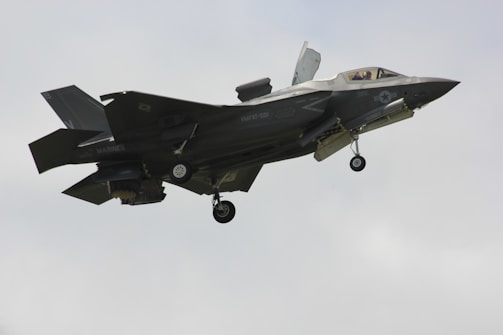 A military fighter jet is captured mid-flight against a pale sky. The aircraft features angular, stealthy design and dark gray color with visible landing gear and some markings on its body.