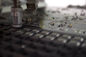 A close-up of a manufacturing machine working on a circuit board. The image focuses on electronic components and a mechanical arm or tool positioned above the board, which is filled with small, intricate parts and traces.