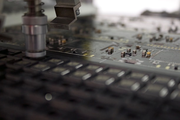 A close-up of a manufacturing machine working on a circuit board. The image focuses on electronic components and a mechanical arm or tool positioned above the board, which is filled with small, intricate parts and traces.