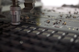 A close-up of a manufacturing machine working on a circuit board. The image focuses on electronic components and a mechanical arm or tool positioned above the board, which is filled with small, intricate parts and traces.