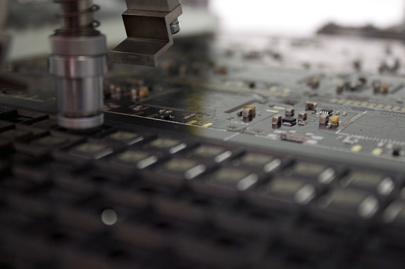 A close-up of a manufacturing machine working on a circuit board. The image focuses on electronic components and a mechanical arm or tool positioned above the board, which is filled with small, intricate parts and traces.