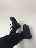 A pair of black combat boots are worn with black leggings. The background is a plain, light-colored wall, highlighting the boots and legs in a relaxed, casual pose.