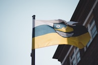 A flag with three horizontal stripes of white, blue, and yellow is waving on a flagpole. The flag features an emblem in the top segment, with a star and a ship against a circular design. It is located near a building with visible windows.