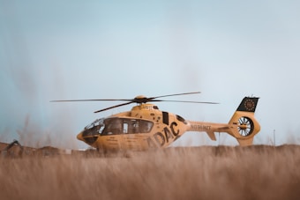A yellow helicopter with the word 'ADAC' and 'NOTARZT' written on its side is parked in a grassy field. The background features a clear sky and some distant structures, giving a sense of an open, rural area.