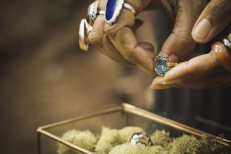 A pair of hands adorned with multiple rings is closely examining a gold ring with a large blue gemstone. The focus is on the detailed rings and the hands, with a blurred background and a glass container filled with green moss in the lower part of the image.