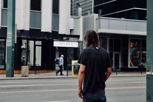 A person with long hair stands on the side of an urban street, looking towards a modern building with large windows and a sign that says 'Avenue.' Another person, wearing a white hat and backpack, walks across the street in the background. The scene suggests a city environment with a mix of pedestrians and traffic infrastructure.