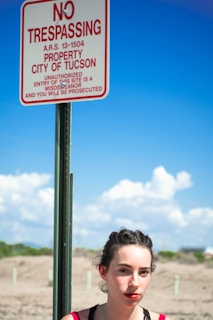 A woman stands beside a 'No Trespassing' sign that displays the message in red on a white background, indicating it's the property of the City of Tucson. The background features a clear blue sky with some fluffy clouds and a dry, desert landscape.