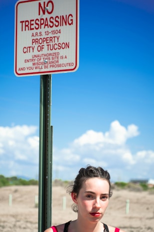 A woman stands beside a 'No Trespassing' sign that displays the message in red on a white background, indicating it's the property of the City of Tucson. The background features a clear blue sky with some fluffy clouds and a dry, desert landscape.