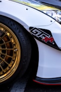 A close-up of a car featuring a shiny gold alloy wheel and a portion of the white bodywork. The wheel has a detailed, intricate design and the tire is black with visible branding. There is a black decal with red text on the side of the car, and the headlight and part of the fender are visible.