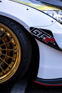 A close-up of a car featuring a shiny gold alloy wheel and a portion of the white bodywork. The wheel has a detailed, intricate design and the tire is black with visible branding. There is a black decal with red text on the side of the car, and the headlight and part of the fender are visible.