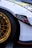 A close-up of a car featuring a shiny gold alloy wheel and a portion of the white bodywork. The wheel has a detailed, intricate design and the tire is black with visible branding. There is a black decal with red text on the side of the car, and the headlight and part of the fender are visible.