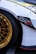 A close-up of a car featuring a shiny gold alloy wheel and a portion of the white bodywork. The wheel has a detailed, intricate design and the tire is black with visible branding. There is a black decal with red text on the side of the car, and the headlight and part of the fender are visible.